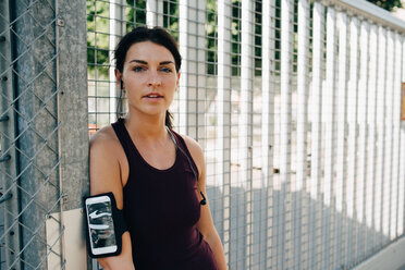 Portrait of confident sportswoman with smart phone and in-ear headphones leaning on railing at bridge - MASF09919