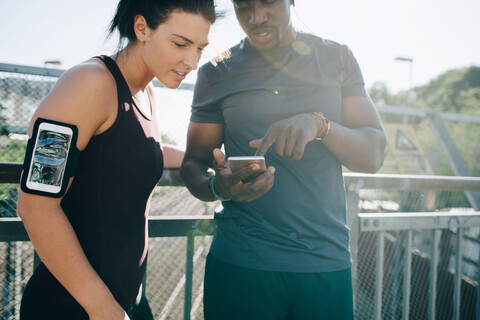 Ein Sportler zeigt einer Sportlerin sein Handy, während er auf einer Brücke am Geländer steht, lizenzfreies Stockfoto