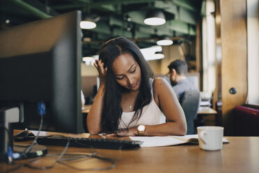 Geschäftsfrau mit Hand im Haar liest Buch am Schreibtisch im Büro - MASF09930