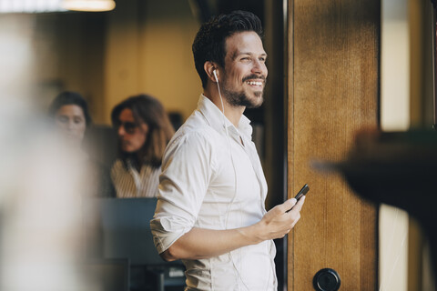 Lächelnder mittelgroßer erwachsener Mann mit Kopfhörern, der mit einem Smartphone im Büro spricht, lizenzfreies Stockfoto