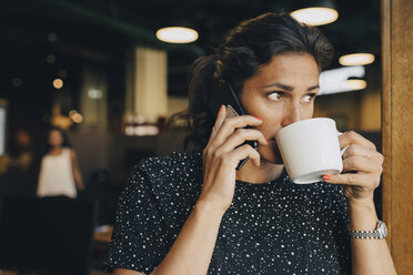 Mittlere erwachsene Geschäftsfrau trinkt Kaffee, während sie ihr Smartphone im Büro benutzt - MASF09944