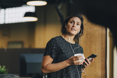 Weibliche Computerprogrammiererin mit Kopfhörern, die mit einem Smartphone im Kreativbüro spricht, lizenzfreies Stockfoto