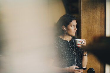 Nachdenkliche Geschäftsfrau, die Kaffee trinkt, während sie im Büro ihr Mobiltelefon benutzt - MASF09946