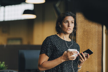 Weibliche Führungskraft hält Kaffeetasse, während sie mit Kopfhörern während eines Telefonats im Büro spricht - MASF09947