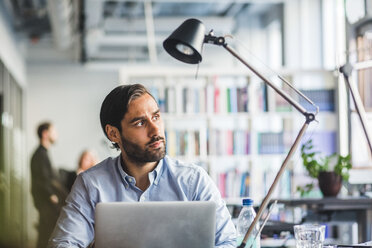 Nachdenklicher Geschäftsmann sitzt mit Laptop am Schreibtisch in einem kreativen Büro - MASF09978