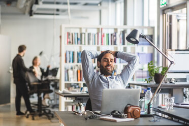 Smiling businessman looking at laptop while sitting with hands behind head in office - MASF09979