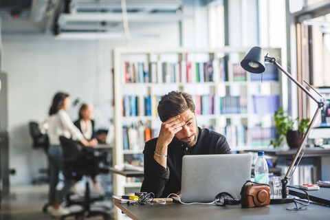 Besorgter männlicher Unternehmer, der auf seinen Laptop schaut, während er im Büro sitzt, lizenzfreies Stockfoto
