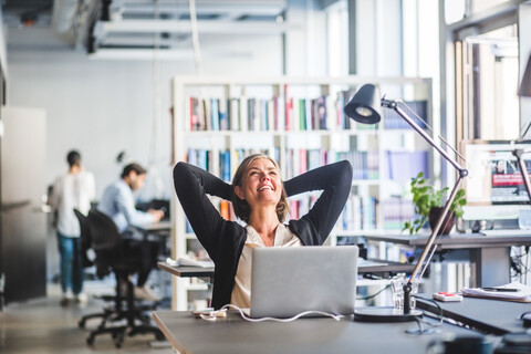 Reife Geschäftsfrau sitzt mit den Händen hinter dem Kopf am Schreibtisch im Büro, lizenzfreies Stockfoto