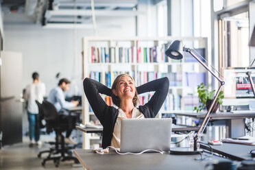 Mature businesswoman sitting with hands behind head at desk in office - MASF09998