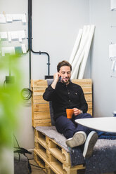 Kreativer Geschäftsmann, der über sein Mobiltelefon spricht, während er auf einem Palettenstapel im Büro sitzt - MASF10023