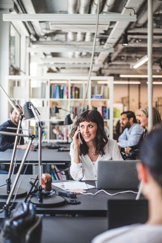 Unternehmerin schaut weg, während sie am Schreibtisch im Kreativbüro mit dem Handy telefoniert, lizenzfreies Stockfoto