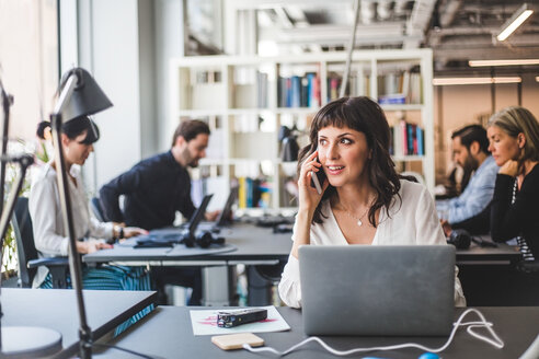 Geschäftsfrau, die wegschaut, während sie am Schreibtisch in einem kreativen Büro mit einem Mobiltelefon spricht - MASF10038