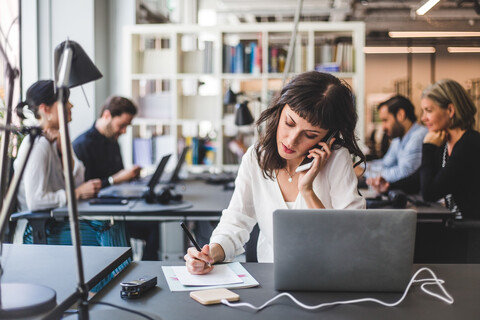 Geschäftsfrau, die am Schreibtisch in einem kreativen Büro mit einem Mobiltelefon spricht, lizenzfreies Stockfoto