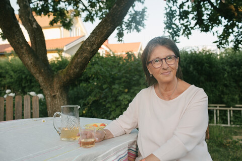 Porträt einer lächelnden Frau, die ein Getränk in der Hand hält, während sie an einem Tisch im Hinterhof sitzt, lizenzfreies Stockfoto