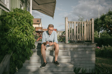 Älterer Mann in voller Länge, der ein Mobiltelefon benutzt, während er auf den Stufen einer Veranda sitzt - MASF10096