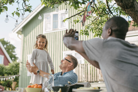 Ein Mann fotografiert seine Tochter, die neben einem älteren Mann am Tisch steht, während einer Gartenparty, lizenzfreies Stockfoto