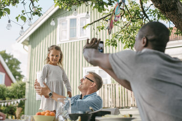 Male photographing daughter standing by senior man at table during garden party - MASF10099