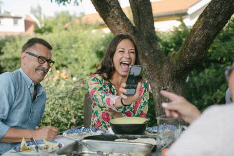 Fröhliche Frau, die einer älteren Frau ihr Smartphone zeigt, während sie bei einer Gartenparty am Tisch sitzt, lizenzfreies Stockfoto