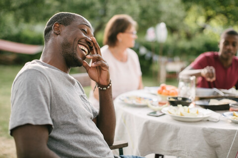 Mittlerer erwachsener Mann lächelt, während er bei einer Gartenparty am Tisch sitzt, lizenzfreies Stockfoto
