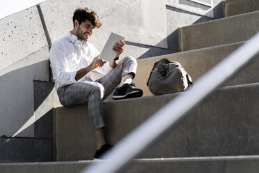 Smiling young man sitting on stairs outdoors using tablet - GIOF04836