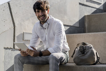 Porträt eines lächelnden jungen Mannes, der auf einer Treppe im Freien sitzt und ein Tablet benutzt - GIOF04839