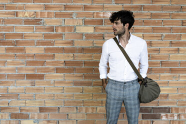 Junger Mann mit Tasche steht an einer Backsteinmauer und schaut sich um - GIOF04855
