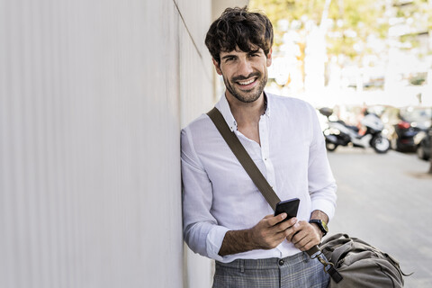 Porträt eines lächelnden jungen Mannes mit Tasche und Mobiltelefon in der Stadt, lizenzfreies Stockfoto