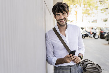 Portrait of smiling young man with bag and cell phone in the city - GIOF04858