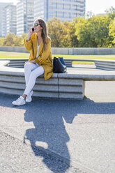 Happy woman with bag and cell phone sitting on a bench in the city - GIOF04871