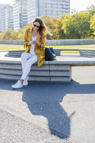 Lächelnde Frau mit Tasche sitzt auf einer Bank in der Stadt und benutzt ein Handy, lizenzfreies Stockfoto