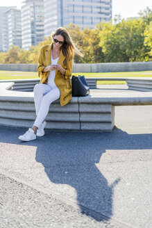 Lächelnde Frau mit Tasche sitzt auf einer Bank in der Stadt und benutzt ein Handy - GIOF04873