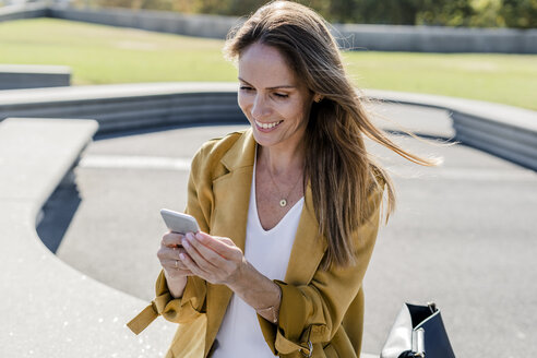 Lächelnde Frau mit Tasche sitzt auf einer Bank in der Stadt und benutzt ein Handy - GIOF04874