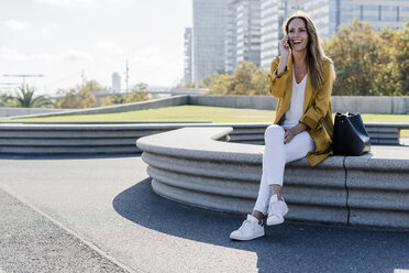 Happy woman with bag and cell phone sitting on a bench in the city - GIOF04876