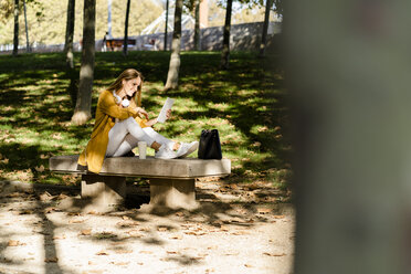 Smiling woman sitting on a bench in a park using tablet - GIOF04892