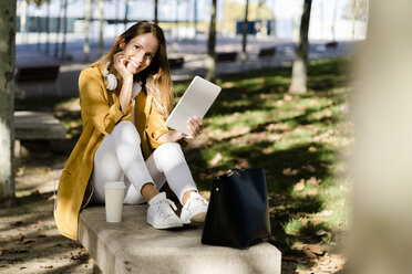 Smiling woman sitting on a bench in a park using tablet - GIOF04894