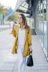 Lächelnde Frau mit Handtasche und Kaffee zum Mitnehmen geht an einem Gebäude entlang - GIOF04898