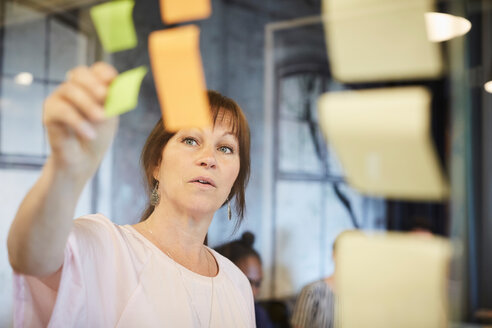 Mature businesswoman sticking sticky note on glass in creative office - MASF10191