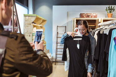 Man photographing smiling woman on smart phone holding dress at store - MASF10208