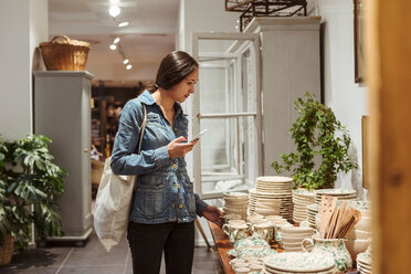 Young woman with mobile phone choosing crockery arranged on table in boutique - MASF10212