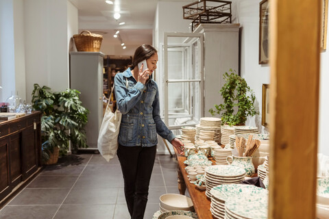 Junge Frau, die mit einem Mobiltelefon spricht und Geschirr auf einem Tisch in einem Geschäft betrachtet, lizenzfreies Stockfoto