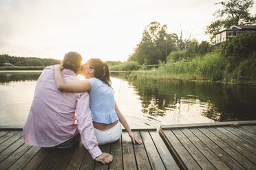 Rückansicht eines Freundes, der seine Freundin küsst, die mit den Armen auf einem Steg über einem See bei Sonnenuntergang sitzt - MASF10264