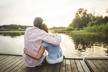 Rear view of couple sitting with arms around on jetty over lake during sunset - MASF10265