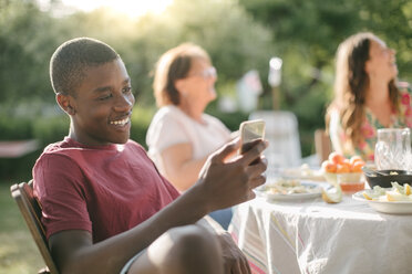 Junge lächelt bei der Benutzung eines Mobiltelefons im Hinterhof während einer Gartenparty - MASF10273