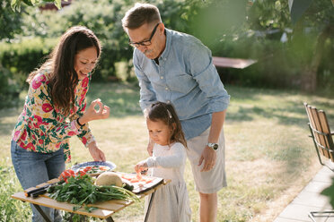 Girl cutting food while standing by mother and grandfather at table in backyard - MASF10283