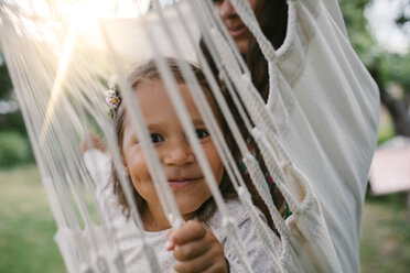 Portrait of smiling girl seen through ropes while swinging with mother on hammock in backyard - MASF10291