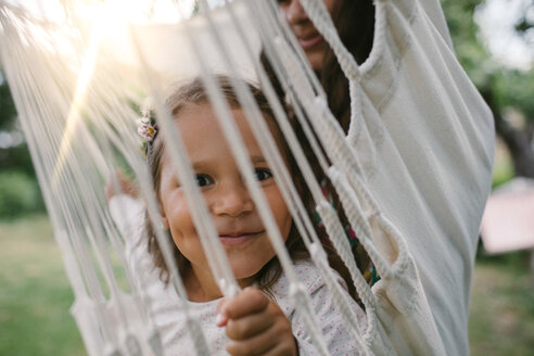 Porträt eines lächelnden Mädchens, das durch Seile gesehen wird, während es mit seiner Mutter in der Hängematte im Garten schaukelt - MASF10291