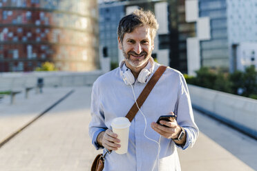 Porträt eines lächelnden reifen Mannes mit Kaffee zum Mitnehmen, Kopfhörern und Mobiltelefon in der Stadt - GIOF04910