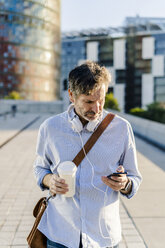 Mature man with takeaway coffee and headphones using cell phone in the city - GIOF04912