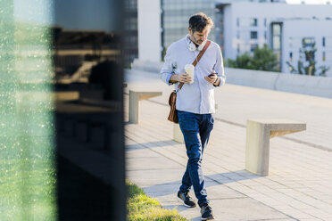 Mature man with takeaway coffee and headphones using cell phone in the city - GIOF04913