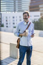 Porträt eines lächelnden reifen Mannes mit Kaffee zum Mitnehmen, Kopfhörern und Mobiltelefon in der Stadt - GIOF04915
